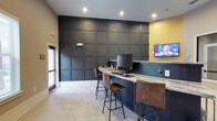 This well-appointed single-floor amenity space blends modern design with inviting functionality. The reception and lounge area welcomes visitors with warm neutral tones, exposed brick accent walls, pendant lighting, and comfortable seating accented with colorful throw pillows. Framed butterfly artwork adds a decorative touch, while a business center workstation tucked nearby provides a practical workspace complete with a desktop computer and printer.

A curved granite-topped bar counter serves as a central hub, surrounded by leather-upholstered bar stools with sleek metal frames. The counter area is equipped with computer workstations and a wall-mounted flat-screen TV, set against a dramatic floor-to-ceiling dark paneled accent wall with a bold geometric grid pattern. A gold-toned accent wall adds a warm contrast to the otherwise cool, sophisticated palette.

The game and recreation area is highlighted by a full-size billiards table bathed in natural light from large bay windows, creating an open and airy atmosphere. Adjacent to this, a dedicated game zone features a shuffleboard table against a dark brick-effect accent wall boldly emblazoned with the phrase "LET'S PLAY!" Throughout the space, large-format light stone tile flooring and herringbone-patterned inlays tie the various zones together cohesively.
