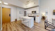 This thoughtfully designed 2-bedroom, 2-bathroom residence offers a clean, modern aesthetic across a single level. The open-concept layout seamlessly connects the kitchen, dining, and living areas, creating an efficient and inviting flow throughout the space. Warm hardwood-style flooring runs continuously through the main living areas, complementing the bright white walls and contemporary finishes.

The kitchen is well-appointed with sleek white cabinetry extending to the ceiling for generous storage, stainless steel appliances including a refrigerator, range, dishwasher, and over-the-range microwave, and a white quartz island with bar seating — ideal for casual dining or entertaining. Track lighting and pendant fixtures add a modern touch overhead.

The living area benefits from floor-to-ceiling windows that draw in abundant natural light and offer urban views. A frosted glass sliding door provides a stylish partition within the main living space. Both bedrooms feature large windows with city views, creating bright and airy retreats.

The two bathrooms are finished in crisp white, with one featuring marble-look tile flooring, a soaking tub, and a single vanity, while the second includes a glass-enclosed walk-in shower. In-unit stacked laundry is also conveniently included. Overall, the space presents a cohesive, modern urban living experience.