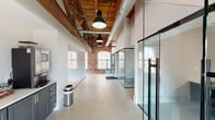 This single-floor commercial office space in South Carolina masterfully marries historic industrial character with contemporary workplace design. The open layout is anchored by soaring ceilings that reveal original heavy timber roof trusses with bold steel hardware, exposed ductwork, and warm wood decking overhead — all set against crisp white walls that amplify the abundant natural light streaming through large arched brick windows.

The polished concrete floors flow throughout, connecting distinct functional zones. Glass-walled private offices and enclosures with sleek black metal framing provide visual transparency while maintaining acoustic separation, giving the space a modern, professional feel without sacrificing its open, airy atmosphere.

A standout conference room features a substantial solid wood boardroom table, executive seating, an original exposed brick accent wall, and a wall-mounted display screen — all framed by the dramatic timber truss ceiling above. The space is further supported by a well-appointed kitchen and break area, finished with slate-blue cabinetry, gold hardware, white countertops, and beverage service equipment.

Industrial pendant lighting punctuates the main corridor, while the preserved brick walls and structural beams throughout create a cohesive aesthetic that honors the building's heritage. This space is ideal for a professional team seeking a distinctive, character-rich work environment.