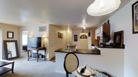 This single-floor residential space offers a thoughtfully designed open-concept layout that seamlessly connects the kitchen, dining, and living areas. The kitchen is a standout feature, centered around a peninsula with a dark countertop and an integrated sink, providing both prep space and a casual dining spot. Dark-finished bar seating complements the peninsula, while warm wood cabinetry lines the walls, offering generous storage. Appliances are neatly integrated, and track lighting overhead ensures the workspace is well-lit. Adjacent to the kitchen, the dining area benefits from a statement pendant light that adds a touch of elegance. The living area is carpeted and comfortably appointed, with space for a media console and television. A floor lamp provides warm ambient lighting, contributing to an inviting atmosphere. The entryway, visible from the main living space, features a closet and connects smoothly to the rest of the unit. Throughout the space, neutral wall tones and consistent ceiling height create a cohesive, airy feel. Wall-mounted decor adds personality without overwhelming the clean aesthetic of the interior.