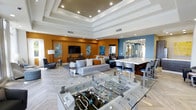 This single-floor amenity lounge is designed with a contemporary aesthetic and a welcoming, social atmosphere. The space features a striking color palette, with warm amber-orange and cool steel-blue accent walls that add energy and visual contrast throughout the open floor plan. A coffered ceiling with recessed lighting adds architectural depth, drawing the eye upward and contributing to an elevated sense of space.

The lounge area is anchored by a plush, light-gray sectional sofa accented with coordinating throw pillows, positioned facing a wall-mounted flat-screen television atop a wood-toned media console. Abstract artwork adorns the walls, reinforcing the modern design sensibility. A glass-top foosball table provides a prominent recreational focal point at the center of the room, making it well-suited for social gatherings.

Adjacent to the lounge, a bar-height counter with sleek white countertops and contemporary bar seating offers a casual dining or socializing area. The light-toned herringbone-patterned flooring ties the zones together cohesively. A mirrored passage in the background suggests additional connected spaces beyond, enhancing the overall sense of openness.