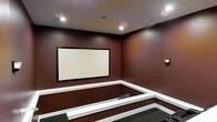 This single-floor space is purpose-built as a private home theater, thoughtfully designed to deliver an immersive entertainment experience. The room is anchored by a large, framed projection screen mounted flush against the front wall, serving as the focal point of the space. Deep burgundy-toned walls envelop the room, creating a rich, cinematic atmosphere that minimizes light reflection and enhances viewing contrast.

The theater features a tiered floor plan, with a raised platform section visible toward the rear of the room, allowing unobstructed sightlines from multiple seating rows. White trim detailing along the platform edges provides a clean visual contrast against the darker walls and carpeted flooring. Recessed ceiling lights illuminate the space efficiently without compromising the theater ambiance, while a wall-mounted sconce adds a subtle accent light along one side wall.

The ceiling is smooth and well-proportioned, contributing to the room's acoustically intentional design. The dark carpet throughout helps absorb sound and reinforces the dedicated media room aesthetic. Overall, this theater space is well-suited for film screenings, gaming, or large-scale media presentations.