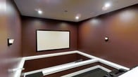 This single-floor space is purpose-built as a private home theater, thoughtfully designed to deliver an immersive entertainment experience. The room is anchored by a large, framed projection screen mounted flush against the front wall, serving as the focal point of the space. Deep burgundy-toned walls envelop the room, creating a rich, cinematic atmosphere that minimizes light reflection and enhances viewing contrast.

The theater features a tiered floor plan, with a raised platform section visible toward the rear of the room, allowing unobstructed sightlines from multiple seating rows. White trim detailing along the platform edges provides a clean visual contrast against the darker walls and carpeted flooring. Recessed ceiling lights illuminate the space efficiently without compromising the theater ambiance, while a wall-mounted sconce adds a subtle accent light along one side wall.

The ceiling is smooth and well-proportioned, contributing to the room's acoustically intentional design. The dark carpet throughout helps absorb sound and reinforces the dedicated media room aesthetic. Overall, this theater space is well-suited for film screenings, gaming, or large-scale media presentations.