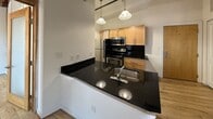 Located at 423 North 18th Street in Richmond, Virginia, this loft-style unit offers a compelling blend of historic industrial character and contemporary design. The space is defined by striking exposed brick walls and original timber ceiling beams, which lend a warm, authentic industrial aesthetic throughout. Light hardwood flooring runs continuously underfoot, complementing the raw architectural elements and contributing to an airy, cohesive feel.

A standout feature is the interior glass partition wall, framed in warm wood, which divides the living area while allowing light to pass freely between spaces. Ceiling fans are mounted to the beamed ceiling, enhancing airflow and adding to the loft's open, relaxed atmosphere. Natural light filters in through windows set into the brick walls, brightening the interior and providing views of the surrounding neighborhood.

The kitchen is well-equipped and functional, featuring dark granite countertops with a peninsula and double-basin sink, warm wood cabinetry, a dark tile backsplash, and stainless steel appliances including a range with microwave above and a refrigerator. Pendant lighting over the kitchen area adds a polished finishing touch. Together, these elements create a space that honors its industrial roots while offering comfortable, modern livability in the heart of Richmond.