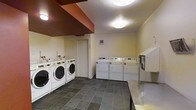 This communal laundry facility is designed with practicality and efficiency in mind, offering residents a well-organized space to handle their laundry needs. The room is outfitted with multiple front-loading washing machines and top-loading dryers arranged along the walls, providing ample capacity for shared use. A stainless steel countertop runs along one side of the room, offering a sturdy and hygienic surface for folding and sorting laundry. The flooring consists of large-format dark slate tiles, which are both durable and easy to maintain, lending the space a clean and utilitarian aesthetic. The walls are finished in a neutral cream tone, keeping the environment bright and open. Overhead, flush-mounted ceiling lights provide consistent, even illumination throughout the room. A payment kiosk is conveniently mounted on the wall near the machines, streamlining the coin or card payment process. Sliding closet doors are also present, offering potential storage access. A wall-mounted ventilation panel ensures proper airflow and climate control within the space. Overall, this laundry room is thoughtfully laid out to maximize usability and accommodate multiple users simultaneously, making it a highly functional shared amenity.
