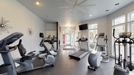 This fitness center is designed to support a comprehensive workout experience within a bright, open environment. The space is organized across a single floor and divided into distinct functional zones, each catering to different aspects of exercise. The cardio area is generously outfitted with multiple treadmills, elliptical trainers, a stationary upright bike, and a recumbent bike, all arranged to maximize floor space and usability. A dedicated strength training room features cable machines, a multi-function weight station, a dumbbell rack, and weight benches, with floor-to-ceiling mirrors lining the walls to aid in proper form. Durable dark rubber flooring spans the workout areas, providing both safety and easy maintenance. The space benefits from abundant natural light streaming through large windows on multiple walls, offering views of the surrounding wooded outdoor scenery. A vaulted ceiling fitted with a large ceiling fan promotes air circulation throughout the main cardio area, while recessed lighting ensures a well-lit environment at all times. A wall-mounted flat-screen television adds an entertainment option for cardio sessions. Near the entrance, hardwood flooring transitions from the workout zones, leading to glass-paneled French doors that open directly onto an outdoor deck. A beverage vending machine and a bulletin board near the entry provide added convenience for users of this well-appointed facility.