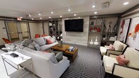This single-floor space showcases a thoughtfully designed living area with a strong contemporary aesthetic. The focal point of the room is a large flat-screen television mounted on a textured stone-panel accent wall, flanked by open built-in shelving units finished in warm grey tones. The shelving provides ample display and storage space, currently styled with decorative vases, books, and curated accessories that reinforce the modern design language.

Recessed lighting is distributed evenly across the ceiling, creating a well-lit, polished atmosphere without relying on natural light sources. The ceiling height contributes to a comfortable, intimate scale throughout the space.

The seating arrangement centers around a sectional sofa and additional accent chairs upholstered in neutral grey tones, accented with coral and teal throw pillows that introduce subtle pops of color. A rich wood-toned coffee table anchors the seating area, resting atop a bold geometric-patterned area rug in grey and white that adds visual depth and texture to the flooring.

Overall, the space presents a cohesive, sophisticated interior with a neutral palette punctuated by deliberate color accents, offering a refined yet inviting environment suited for both relaxation and entertainment.
