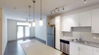 This single-floor space showcases a clean, contemporary design centered around an open-concept layout that seamlessly connects the kitchen and living areas. The kitchen is appointed with floor-to-ceiling white cabinetry, offering generous storage, while stainless steel appliances — including a full-size refrigerator and dishwasher — add a polished, modern touch. A light-toned countertop extends along the kitchen island, providing an ideal surface for meal prep or casual dining. Gray subway tile backsplash adds subtle texture and visual contrast against the bright white cabinetry. Overhead, a track lighting system with adjustable spotlights ensures the kitchen is well-lit, while a trio of cylindrical pendant lights hangs above the island, contributing both function and modern style. The adjacent living area benefits from natural light streaming through large windows with horizontal blinds, creating a bright and welcoming atmosphere. White walls and neutral flooring throughout maintain a cohesive, airy feel, making the space feel open and well-proportioned. The overall ambiance is modern and minimalist, with thoughtful lighting and clean lines defining every corner.
