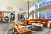 Located in Atlanta, GA, this single-floor community lounge is a masterclass in contemporary design, combining elevated materials and thoughtful layouts to create an inviting social hub. The space is anchored by a generous central bar island finished in striking white marble, topped with sculptural geometric pendant lights and backed by a textured stone tile backsplash with open shelving and built-in beverage stations. Dark-stained wood cabinetry adds warmth and contrast throughout.

Two distinct seating areas define the lounge. The first features a dramatic double-height ceiling, floor-to-ceiling windows flooding the room with natural light, and a dark slatted wood accent wall hosting a wall-mounted television. Cognac leather sofas and armchairs are arranged atop a layered area rug, creating an inviting gathering spot. The second seating area showcases a geometric patterned wood accent wall with brass globe sconces, complemented by a tufted leather banquette and modern side tables.

Large-format tile flooring flows throughout, tying the areas together cohesively. A corner section features wraparound floor-to-ceiling glazing that frames serene courtyard views of a pool and landscaped grounds. Curated artwork adorns the crisp white walls, reinforcing the space's refined, contemporary character.