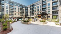 Located at 100 Waddell Plaza Ln in Cary, NC, this thoughtfully designed outdoor amenity courtyard is enclosed by a striking multi-story building featuring a contemporary mix of brick, glass, and modern façade elements. The centerpiece of the space is a resort-style swimming pool with a gently curving shape, complemented by in-pool lounge chairs and a decorative pergola-style feature that adds a distinctive architectural touch. The pool area is enclosed by sleek iron fencing and surrounded by well-maintained landscaped beds with seasonal plantings and manicured shrubbery.

Adjacent to the pool, a dedicated putting green offers a multi-hole layout set into lush synthetic turf, providing a recreational outlet set against the backdrop of the building's curved facade. Outdoor seating areas are thoughtfully distributed throughout, including a covered pergola lounge with string lighting, upholstered sectional and accent chairs in a navy palette, and a wall-mounted display screen — ideal for social gatherings. Additional patio dining areas featuring blue round tables and woven chairs are positioned throughout the courtyard's paved sections. Raised planter beds with flowering plants and ground-level greenery add natural color and texture, creating a vibrant, resort-like ambiance in the heart of Cary, NC.