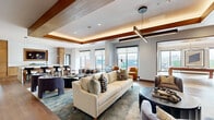 Located in Oakland's 94608 zip code, this thoughtfully designed single-floor amenity space blends contemporary architecture with warm, inviting interiors. The open-plan layout flows seamlessly between multiple distinct zones, unified by rich hardwood flooring, coffered ceilings with warm wood trim, and abundant natural light streaming through large floor-to-ceiling windows that frame urban street views.

The main lounge area anchors the space, featuring a tray ceiling with recessed LED uplighting and a striking linear pendant fixture. Plush seating arrangements—including cream sofas, velvet armchairs in slate and cognac tones, and layered area rugs—create a welcoming social environment. An adjacent bar area with marble countertops, a wine refrigerator, integrated sink, and warm wood cabinetry adds a refined hospitality element.

A dedicated reception-style lounge area showcases a teal accent wall with curated artwork, swivel accent chairs, and built-in banquette seating. The billiards room offers a full-size pool table beneath a sculptural polished-chrome chandelier, surrounded by windows with open city views. Throughout the space, the consistent palette of warm woods, soft neutrals, and teal accents creates a cohesive, upscale ambiance.