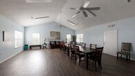Located at 8242 Durelee Ln in Douglasville, GA, this single-floor clubhouse offers a well-appointed and versatile gathering space designed to accommodate community events and social functions. The main hall is defined by striking vaulted ceilings with dual ceiling fans that enhance airflow and add architectural character. Light blue walls throughout create a calm, welcoming atmosphere, while dark hardwood-style flooring provides a rich contrast and a polished finish underfoot. Large windows fitted with white blinds line the walls, filling the space with abundant natural light. The open-concept layout seamlessly integrates a generous dining area — anchored by a long dark wood table and ample seating — with a comfortable lounge section featuring a brown sofa, accent chairs, and decorative furnishings. A dedicated kitchen area offers dark cabinetry, granite-style countertops, a double stainless steel sink, and a window that brings in additional daylight, making it fully functional for catered events or community use. A hallway provides access to additional areas of the building. The space also includes a wall-mounted display area and a TV stand, adding media capability to the room. Overall, this clubhouse delivers a bright, spacious, and functional environment suited for a wide range of community activities.