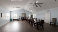 Located at 8242 Durelee Ln in Douglasville, GA, this single-floor clubhouse offers a well-appointed and versatile gathering space designed to accommodate community events and social functions. The main hall is defined by striking vaulted ceilings with dual ceiling fans that enhance airflow and add architectural character. Light blue walls throughout create a calm, welcoming atmosphere, while dark hardwood-style flooring provides a rich contrast and a polished finish underfoot. Large windows fitted with white blinds line the walls, filling the space with abundant natural light. The open-concept layout seamlessly integrates a generous dining area — anchored by a long dark wood table and ample seating — with a comfortable lounge section featuring a brown sofa, accent chairs, and decorative furnishings. A dedicated kitchen area offers dark cabinetry, granite-style countertops, a double stainless steel sink, and a window that brings in additional daylight, making it fully functional for catered events or community use. A hallway provides access to additional areas of the building. The space also includes a wall-mounted display area and a TV stand, adding media capability to the room. Overall, this clubhouse delivers a bright, spacious, and functional environment suited for a wide range of community activities.