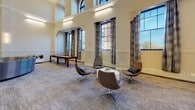 The Great Room is a generously proportioned communal space defined by its dramatic soaring ceilings adorned with detailed crown molding and arched transom windows that flood the interior with natural light. The room is thoughtfully organized into several distinct activity and lounge zones, all unified by neutral-toned carpet flooring and a warm, welcoming palette of cream and soft gray walls accented with wainscoting.

A central high-top communal table with teal upholstered barstools serves as a social hub, while nearby clusters of round café tables paired with deep plum chairs invite smaller gatherings. A curved sectional sofa anchors a relaxed lounge area, complemented by swivel accent chairs and side tables. A wall-mounted flat-screen TV flanked by sleek sconce lighting creates a dedicated viewing nook with comfortable lounge seating.

A stylish built-in kitchenette features warm wood cabinetry, a textured geometric tile backsplash, a sink, microwave, and beverage cooler. Recreation is well-served with a foosball table and a shuffleboard table. Curated abstract artwork is displayed throughout, adding vibrant color and artistic character. Potted greenery, a decorative credenza, and elegant chandelier accents complete the refined yet inviting ambiance of this multifunctional space.