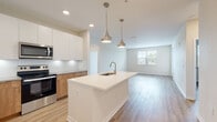 This well-appointed single-floor residence showcases a contemporary design with a cohesive, light-toned palette and light hardwood flooring that flows seamlessly throughout. The open-concept kitchen is a standout feature, centered around a large island with a white quartz countertop and an undermount sink, illuminated by a pair of elegant brushed nickel pendant lights. The kitchen is outfitted with two-tone cabinetry — crisp white upper cabinets paired with warm wood lower cabinets — complemented by a classic subway tile backsplash and a full suite of stainless steel appliances, including a refrigerator with ice dispenser, electric range, and over-the-range microwave. The kitchen opens directly into a spacious living area filled with natural light from multiple windows with views to the outdoors. The bedrooms are generously sized, each benefiting from natural light, while the primary bathroom features a tiled walk-in shower with a glass enclosure and access to a walk-in closet. A second bathroom includes a full soaking tub with a tiled surround and a warm wood vanity. Recessed lighting and clean white walls throughout contribute to the bright, modern ambiance of this thoughtfully designed space.
