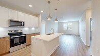 This well-appointed single-floor residence showcases a contemporary design with a cohesive, light-toned palette and light hardwood flooring that flows seamlessly throughout. The open-concept kitchen is a standout feature, centered around a large island with a white quartz countertop and an undermount sink, illuminated by a pair of elegant brushed nickel pendant lights. The kitchen is outfitted with two-tone cabinetry — crisp white upper cabinets paired with warm wood lower cabinets — complemented by a classic subway tile backsplash and a full suite of stainless steel appliances, including a refrigerator with ice dispenser, electric range, and over-the-range microwave. The kitchen opens directly into a spacious living area filled with natural light from multiple windows with views to the outdoors. The bedrooms are generously sized, each benefiting from natural light, while the primary bathroom features a tiled walk-in shower with a glass enclosure and access to a walk-in closet. A second bathroom includes a full soaking tub with a tiled surround and a warm wood vanity. Recessed lighting and clean white walls throughout contribute to the bright, modern ambiance of this thoughtfully designed space.