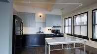 This single-floor space showcases a thoughtfully designed interior that blends industrial character with modern finishes. The kitchen serves as a central highlight, featuring a striking two-tone cabinet design with light gray upper cabinets and dark matte lower cabinets that create a bold contrast. A decorative scalloped white tile backsplash adds texture and visual interest, complementing the stainless steel gas range and built-in range hood overhead. A freestanding island with a white marble top and metal frame provides additional workspace and a refined focal point in the room. Polished concrete flooring throughout reinforces the industrial aesthetic, while exposed ceiling beams and track lighting add to the loft-like ambiance. Large windows with slatted blinds allow natural light to filter into the space, enhancing the open and airy feel. The overall design strikes a balance between raw, urban elements and clean, contemporary finishes, resulting in a space that is both functional and visually compelling. The cohesive material palette and attention to detail make this a distinctive and well-appointed living environment.