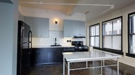 This single-floor space showcases a thoughtfully designed interior that blends industrial character with modern finishes. The kitchen serves as a central highlight, featuring a striking two-tone cabinet design with light gray upper cabinets and dark matte lower cabinets that create a bold contrast. A decorative scalloped white tile backsplash adds texture and visual interest, complementing the stainless steel gas range and built-in range hood overhead. A freestanding island with a white marble top and metal frame provides additional workspace and a refined focal point in the room. Polished concrete flooring throughout reinforces the industrial aesthetic, while exposed ceiling beams and track lighting add to the loft-like ambiance. Large windows with slatted blinds allow natural light to filter into the space, enhancing the open and airy feel. The overall design strikes a balance between raw, urban elements and clean, contemporary finishes, resulting in a space that is both functional and visually compelling. The cohesive material palette and attention to detail make this a distinctive and well-appointed living environment.