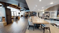 This single-floor space is thoughtfully designed to serve as a multifunctional gathering and work environment. At its center, a long, light wood communal table flanked by upholstered cream chairs provides an ideal setting for collaborative work or casual meetings. The open-plan layout seamlessly transitions into a lounge area furnished with plush armchairs and ambient pendant lighting, creating a relaxed retreat within the same space.

Wide-plank hardwood flooring runs throughout, grounding the interior with warmth and continuity. The ceiling design is notably dynamic, contrasting dark, exposed structural elements and recessed lighting against crisp white sections, adding visual depth and a contemporary industrial edge. A decorative open-frame room divider with circular motifs elegantly separates zones while maintaining an airy, connected feel.

Floor-to-ceiling windows along the perimeter flood the space with natural light and offer views of surrounding greenery. Additional bar-height seating and intimate nook areas further enhance the versatility of the layout. The overall ambiance is polished yet approachable, balancing modern design with comfort.