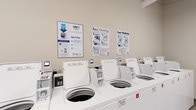 This single-floor amenity space is designed to support the everyday needs of its users, offering two distinct and well-maintained areas. The centerpiece is a generously sized fitness center that makes a strong impression with its open layout, rubber-floored exercise area, and abundant natural light streaming through large windows. The gym is outfitted with a comprehensive selection of TRUE-brand cardio machines, including treadmalls and ellipticals, as well as cable strength-training stations, rowing machines, and free weights. Full-length wall mirrors expand the sense of space and support functional training, while bright linear LED lighting illuminates the entire area. Exposed ceiling infrastructure adds an industrial character, and a raised platform area with stair access contributes to the dynamic layout of the room. Wall-mounted flat-screen televisions enhance the cardio experience. A clean, well-lit corridor connects the fitness center to the adjacent laundry facility, which is equipped with a row of top-load washing machines operated through the CSC payment system, accepting both card and mobile payments. Instructional signage is posted throughout the laundry room for user convenience. Together, these amenities offer a practical and functional environment designed for daily use.