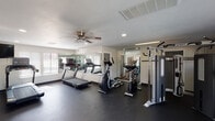 Located at 3012 Ordway Dr NW in Roanoke, VA, this fitness center is a well-equipped, single-floor facility designed to support a comprehensive workout experience. The space features durable rubber flooring throughout, providing a safe and functional surface suited for a variety of exercise activities.

The cardio area is outfitted with multiple treadmills and elliptical machines, allowing users to engage in sustained aerobic training. Full-wall mirrors line portions of the space, visually expanding the room while enabling users to monitor their form. Ceiling fans are installed overhead, complementing the recessed lighting and HVAC vents to maintain a comfortable climate during workouts.

The strength training section includes a multi-function cable machine and an adjustable weight bench, offering options for resistance and functional training. A wall-mounted television is positioned for visibility from the cardio equipment, providing entertainment during sessions.

Natural light enters through several windows fitted with horizontal blinds, contributing to an open and airy atmosphere. The entrance features a set of double doors with glass panels, alongside a secondary interior door providing access to additional areas. The overall design is clean and functional, with white walls and a neutral color palette creating a focused and distraction-free environment.
