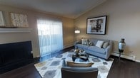 This single-floor space showcases a thoughtfully designed living room that balances comfort and modern style. The room features rich, dark hardwood flooring that contrasts elegantly with the warm, neutral-toned walls, creating a welcoming and cohesive aesthetic throughout.

A standout feature of the living area is the expansive sliding patio door dressed with vertical blinds, allowing an abundance of natural light to fill the room while providing direct access to an outdoor space. A baseboard heater runs beneath the window, ensuring year-round comfort.

The living room is furnished with a contemporary grey sofa accented by decorative pillows in neutral and gold tones, complemented by a matching armchair with a warm-toned throw. A glass-topped oval coffee table with a sculptural metallic base serves as a focal point, anchored by a geometric-patterned area rug in grey, white, and gold. A sleek floor lamp provides additional ambient lighting, while a large framed piece of wall art adds a refined artistic touch to the space. The overall ambiance is clean, modern, and inviting.