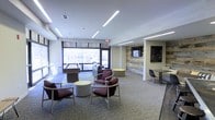 This single-floor communal space is thoughtfully designed to serve as a versatile gathering and recreation area. The open-plan layout is anchored by a series of sleek, linear LED ceiling fixtures that cast consistent, even illumination throughout the room, complementing the generous natural light that pours in through large floor-to-ceiling windows along the exterior wall.

The seating area features a collection of modern armchairs in deep burgundy tones paired with round accent tables, creating an inviting arrangement for casual conversation or relaxation. A reclaimed wood accent wall serves as a striking focal point, with a wall-mounted flat-screen television integrated seamlessly into its surface, adding warmth and texture to the otherwise contemporary design palette.

Toward the windowed perimeter, a billiards table provides a recreational focal point, while bar-height seating along the windows takes advantage of the outdoor views. A dedicated work or dining area with a narrow table and contemporary chairs occupies one side of the space, offering a quieter zone for focused use.

The flooring throughout features a neutral, patterned carpet that ties the various zones together cohesively. The overall ambiance balances modern aesthetics with comfortable, functional design — well-suited for community use, collaboration, or leisure.