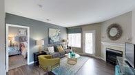 This single-floor space presents a well-appointed living area that blends contemporary design with a warm, inviting ambiance. The living room showcases rich hardwood-style flooring that grounds the space, complemented by neutral-toned walls in soft grey and taupe that create a calm, cohesive aesthetic throughout.

The room is anchored by a comfortable grey sofa adorned with patterned and mustard-yellow accent pillows, paired with a bold chartreuse accent chair that introduces a lively pop of color. A round glass-and-gold coffee table serves as a focal point, sitting atop a soft teal area rug that adds texture and depth to the space.

Natural light flows generously into the room through a large window fitted with white horizontal blinds and a teal fabric valance, while a glass-panel door provides direct access to an outdoor area, further enhancing the brightness of the interior. The walls are decorated with an expressive abstract canvas in teal, grey, and gold tones, tying the color palette together cohesively.

Ambient lighting is provided by stylish table lamps positioned at either end of the sofa, adding warmth during evening hours. A classic white fireplace mantel is partially visible, hinting at additional architectural character within the space.