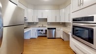This single-floor unit at Rolling Green in Amherst, Massachusetts, is thoughtfully designed with accessibility and functionality at the forefront. The layout, visible in the floor plan, flows logically between living, sleeping, and utility areas, all connected by warm hardwood-style flooring that runs consistently throughout the space.

The open living area is bright and spacious, featuring large windows with vertical blinds that allow natural light to fill the room. A wall-mounted air conditioning unit and baseboard heating ensure year-round comfort. An electrical panel is conveniently integrated into the living space.

The kitchen is well-appointed with white shaker-style cabinetry extending to the ceiling for ample storage, light-toned countertops, and a full suite of stainless steel appliances including a refrigerator, dishwasher, wall oven, and microwave. The under-counter clearance reflects an accessibility-conscious design.

The bedrooms feature hardwood-style floors, natural light from windows, and generous closet space, including a large double bifold closet. A dedicated laundry closet with an in-unit washer is also present.

The bathroom is fully ADA-compliant, offering a roll-in shower with grab bars, a handheld showerhead, a wall-mounted sink, and wide clearances throughout, prioritizing safe and comfortable use.