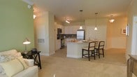 This single-floor residence showcases an open-concept layout that connects the kitchen and living areas into one cohesive, functional space. The kitchen is a standout feature, designed with white cabinetry that extends to the ceiling, a dark tile backsplash, and a full suite of stainless steel appliances including a refrigerator, electric range, and built-in microwave. A central kitchen island serves as both a prep space and casual dining area, accommodated by dark-framed upholstered counter-height chairs. Two elegant pendant lights hang above the island, providing focused task lighting that complements the track lighting overhead. The flooring transitions from tile in the kitchen to plush neutral carpeting in the living area, subtly defining each zone while maintaining visual continuity. Walls painted in soft green and warm beige tones contribute to an inviting atmosphere. The living area offers generous open space, with a small accent table visible near the edge of the room. Overall, the space delivers a practical yet comfortable layout with a clean, well-maintained aesthetic.