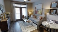 This single-story space showcases a thoughtfully designed living area that balances comfort and modern style. The room is anchored by a light gray sofa adorned with decorative pillows in warm rust and neutral tones, complemented by a geometric-patterned area rug in white and gray that defines the seating zone. A sleek glass-and-metal coffee table serves as the centerpiece, adding an airy, open feel to the space.

Dark hardwood-style flooring runs throughout, providing a rich contrast against the soft gray walls. A large piece of colorful artwork above the sofa adds a vibrant focal point to the room. Natural light flows generously through full-height glass French doors fitted with white curtains and horizontal blinds, which open to a private balcony — extending the living space outdoors. A ceiling fan overhead ensures year-round comfort.

Subtle layering of ambient lighting from table lamps enhances the warm, inviting atmosphere. A small dining area is partially visible, suggesting a practical, open layout. Overall, the space presents a clean, contemporary aesthetic with cohesive design choices throughout.
