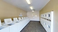This shared laundry facility is a practical, single-floor space designed to accommodate high-volume laundry needs. The room is laid out efficiently, with a long row of top-loading washing machines lining one wall, providing ample capacity for multiple users at once. Along the opposite wall, stacked dryer units are arranged to maximize the available floor space while keeping the overall layout open and easy to navigate. The flooring features wood-look vinyl planking in a medium-dark tone, lending a clean and modern feel to the utilitarian space. Overhead fluorescent lighting ensures the room is well-lit throughout. The walls are finished in a warm neutral tone, contributing to a bright and welcoming atmosphere. A payment or information panel is mounted on the wall, along with posted notices and instructional signage to assist users. The ceiling includes a ventilation duct, supporting proper airflow throughout the facility. Overall, the space is thoughtfully organized, clean, and functional, offering residents a straightforward and accessible laundry experience.