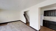 This two-floor space offers a clean, straightforward layout designed for practical everyday living. The main level features a spacious carpeted living area with neutral walls and dark baseboards that provide a subtle contrast throughout. A textured ceiling adds a traditional architectural touch overhead. An open staircase with a dark wood handrail leads to the upper floor, conveniently positioned within the main living space for easy access between levels. Adjacent to the living area, the kitchen is separated by a partial wall, offering a degree of definition while maintaining an open feel. The kitchen is equipped with a white refrigerator and a dark appliance, set against hard flooring that contrasts with the carpeted living zone. The overall color palette is neutral and understated, with white walls and light tones creating a bright, airy atmosphere throughout. The layout maximizes usable space across both floors, making it well-suited for a variety of configurations. The clean lines and simple finishes give the space a versatile foundation that can be easily adapted to suit different needs and styles.
