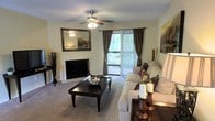 This single-story living space offers a comfortable and well-appointed interior centered around a white-brick fireplace, which serves as the room's focal point. A large framed mirror mounted above the mantle enhances the sense of depth and reflects natural light throughout the space. The living area is finished with plush wall-to-wall carpeting in a warm neutral tone, contributing to the overall inviting atmosphere. A ceiling fan with an integrated light fixture is centrally positioned, providing both airflow and ambient lighting. The room receives generous natural light through a sliding glass door dressed with floor-length, dark drape panels and vertical blinds, offering a view of lush greenery outside. The layout comfortably accommodates a generous sectional sofa arrangement paired with a dark wood coffee table, creating a practical yet stylish seating area. Decorative accents, including candles and botanical arrangements, add warmth to the space. The textured ceiling and soft, neutral wall color tie the room together, creating a cohesive and relaxed aesthetic suited for everyday living.