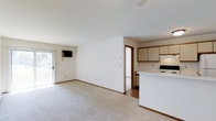 This single-floor space presents an open-concept layout that seamlessly integrates the living area and kitchen into one cohesive environment. The living area is finished with wall-to-wall carpet in a neutral tone, contributing to a warm and understated ambiance. A ceiling fan is positioned overhead, providing both ventilation and a subtle design element. A wall-mounted television and a sliding glass door that introduces natural light and offers a view to the outdoors are notable features of the living area.

The kitchen is adjacent to the living space, separated by a raised peninsula that serves as both a functional workspace and a visual divider. Light-toned cabinetry with wood-trimmed accents lines the upper portion of the kitchen walls, providing ample storage. A white electric range and additional appliances are integrated into the kitchen layout. The kitchen flooring transitions from the carpeted living area to a hard surface, adding a practical distinction between the two zones. Smooth white walls and consistent trim throughout the space reinforce a clean, cohesive aesthetic.
