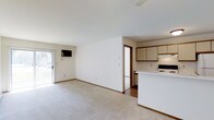 This single-floor space presents an open-concept layout that seamlessly integrates the living area and kitchen into one cohesive environment. The living area is finished with wall-to-wall carpet in a neutral tone, contributing to a warm and understated ambiance. A ceiling fan is positioned overhead, providing both ventilation and a subtle design element. A wall-mounted television and a sliding glass door that introduces natural light and offers a view to the outdoors are notable features of the living area.

The kitchen is adjacent to the living space, separated by a raised peninsula that serves as both a functional workspace and a visual divider. Light-toned cabinetry with wood-trimmed accents lines the upper portion of the kitchen walls, providing ample storage. A white electric range and additional appliances are integrated into the kitchen layout. The kitchen flooring transitions from the carpeted living area to a hard surface, adding a practical distinction between the two zones. Smooth white walls and consistent trim throughout the space reinforce a clean, cohesive aesthetic.