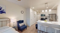 This contemporary single-floor unit offers a well-designed open-concept layout that seamlessly integrates the living, dining, and kitchen areas. The space is finished with light hardwood-style flooring throughout, recessed lighting, and neutral white walls that lend a clean, bright aesthetic.

The kitchen is a standout feature, outfitted with white shaker-style cabinetry, stainless steel appliances including a refrigerator, electric range, and over-the-range microwave, as well as a white quartz peninsula with an undermount sink. Pendant lighting above the peninsula adds a modern touch, and bar-style seating makes it ideal for casual dining or entertaining.

The living area is an intimate, cozy space that flows naturally from the kitchen, offering room for seating and relaxation. A hallway connects the main living space to the private bedroom and bathroom.

The bedroom is a serene retreat with a ceiling fan, a wall-mounted television, and a large window that frames lush greenery outside, filling the room with natural light and a calming outdoor view.

The bathroom is finished with warm neutral tones, a white vanity with ample cabinet storage, a large mirror, and a shower-tub combination, keeping the space functional and tidy. In-unit laundry is also visible, adding everyday convenience to this thoughtfully designed home.
