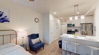This contemporary single-floor unit offers a well-designed open-concept layout that seamlessly integrates the living, dining, and kitchen areas. The space is finished with light hardwood-style flooring throughout, recessed lighting, and neutral white walls that lend a clean, bright aesthetic.

The kitchen is a standout feature, outfitted with white shaker-style cabinetry, stainless steel appliances including a refrigerator, electric range, and over-the-range microwave, as well as a white quartz peninsula with an undermount sink. Pendant lighting above the peninsula adds a modern touch, and bar-style seating makes it ideal for casual dining or entertaining.

The living area is an intimate, cozy space that flows naturally from the kitchen, offering room for seating and relaxation. A hallway connects the main living space to the private bedroom and bathroom.

The bedroom is a serene retreat with a ceiling fan, a wall-mounted television, and a large window that frames lush greenery outside, filling the room with natural light and a calming outdoor view.

The bathroom is finished with warm neutral tones, a white vanity with ample cabinet storage, a large mirror, and a shower-tub combination, keeping the space functional and tidy. In-unit laundry is also visible, adding everyday convenience to this thoughtfully designed home.