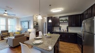 This single-floor residence offers a thoughtfully designed open layout that seamlessly connects the kitchen, dining, and living areas. The kitchen is a standout feature, showcasing rich dark espresso cabinetry reaching toward the ceiling, granite countertops, a white subway tile backsplash, and a full suite of stainless steel appliances including a range, microwave, dishwasher, and refrigerator. A granite-topped island provides additional prep space and casual dining seating illuminated by pendant lighting overhead. Warm hardwood-style flooring flows throughout the main living areas, enhancing the cohesive aesthetic. The living room is bright and welcoming, with large windows allowing generous natural light and access to an exterior door. Two bedrooms offer comfortable retreats, each featuring plush carpeting and neutral toned walls. The bathrooms are finished with dark vanity cabinetry, granite countertops, and hardwood-style flooring. One bathroom features a tub-shower combination with tile surround, while the second offers a spacious layout with ample room. Throughout the residence, a consistent gray and neutral color palette, accented by warm wood tones, creates a modern and inviting ambiance.