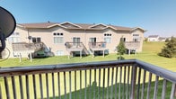 This single-storey residence is part of a row-style multi-unit complex, constructed with light-toned vinyl siding beneath a pitched, dark shingled roofline. The building's exterior design is characterized by repeating gabled sections, giving each unit a distinct architectural identity while maintaining a cohesive community aesthetic. Each unit features its own raised wooden deck, accessible from the interior and supported by posts above a ground-level storage or utility space. The decks are outfitted with railings and offer views of a shared, well-kept grassy courtyard that runs along the back of the building. The outdoor area provides an open, communal green space with mature trees interspersed throughout, enhancing the natural ambiance. Windows along the rear facade allow for natural light and outward views of the landscaped grounds. The elevated deck position offers a pleasant vantage point over the lawn, making it a practical and enjoyable outdoor extension of the living space. The overall exterior presents a tidy, community-oriented environment well-suited for everyday living.