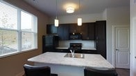 This single-floor space showcases a well-appointed kitchen as its centerpiece, designed with a cohesive and contemporary aesthetic. The kitchen is outfitted with rich, dark espresso-toned cabinetry that extends from base to upper wall units, providing generous storage throughout. The cabinetry is paired with light-toned granite countertops featuring subtle veining, creating a striking contrast that anchors the room's design. A large kitchen island with a built-in stainless steel sink serves as both a functional workspace and a casual dining area, accommodated by bar-height seating. Black appliances, including a refrigerator, gas range, and over-the-range microwave, complement the dark cabinetry seamlessly. Overhead, a combination of pendant lights above the island and a flush-mount ceiling fixture provides layered lighting that enhances both task and ambient illumination. A window fitted with horizontal blinds allows natural light to enter the space, brightening the kitchen and adding to its open, inviting atmosphere. The overall design is clean and modern, with thoughtful material choices that create a polished, functional environment.