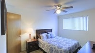 This single-floor space showcases a well-appointed bedroom designed for comfort and functionality. The room features smooth, neutral-toned walls that contribute to a calm and restful ambiance. A ceiling fan with an integrated light fixture is centrally mounted overhead, providing both air circulation and ambient lighting throughout the space. The room receives natural light through a window dressed with vertical blinds, allowing for adjustable light control and privacy. The flooring appears to be a low-pile carpet or smooth surface, adding to the room's understated, practical design. The overall layout is efficiently organized, offering enough space to comfortably accommodate a standard bed, nightstand, and additional furnishings. The warm glow from a bedside lamp complements the natural light, creating a layered lighting scheme suited for relaxation. The dark-wood nightstand with drawer storage adds a functional and visually grounding element to the space. Overall, the room presents a straightforward, comfortable living environment with thoughtful details that balance practicality and coziness, making it a welcoming personal retreat.