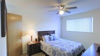 This single-floor space showcases a well-appointed bedroom designed for comfort and functionality. The room features smooth, neutral-toned walls that contribute to a calm and restful ambiance. A ceiling fan with an integrated light fixture is centrally mounted overhead, providing both air circulation and ambient lighting throughout the space. The room receives natural light through a window dressed with vertical blinds, allowing for adjustable light control and privacy. The flooring appears to be a low-pile carpet or smooth surface, adding to the room's understated, practical design. The overall layout is efficiently organized, offering enough space to comfortably accommodate a standard bed, nightstand, and additional furnishings. The warm glow from a bedside lamp complements the natural light, creating a layered lighting scheme suited for relaxation. The dark-wood nightstand with drawer storage adds a functional and visually grounding element to the space. Overall, the room presents a straightforward, comfortable living environment with thoughtful details that balance practicality and coziness, making it a welcoming personal retreat.