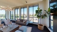 This single-floor amenity space is designed with a contemporary aesthetic and offers an impressive array of features suited for both relaxation and collaboration. The layout encompasses a generous open lounge area and a dedicated conference or meeting room, each thoughtfully appointed and bathed in natural light.

The lounge area serves as the centerpiece of the space, anchored by a sleek, dark-framed fireplace that adds warmth and visual drama. A sculptural antler-style chandelier overhead complements the modern furnishings, which include a plush sectional sofa, striped coffee table, and soft ottomans arranged atop a layered area rug. Floor-to-ceiling windows wrap the lounge, framing sweeping views of the surrounding cityscape and treetops, while access to an outdoor terrace extends the living space further.

The conference area is equally well-lit, featuring large-format windows on multiple walls that flood the room with daylight and offer expansive views of the urban landscape and green canopy below. A solid wood conference table paired with modern white chairs creates a clean, professional environment ideal for meetings or focused work.

Throughout the space, dark carpet flooring, neutral wall tones, and bold window framing create a cohesive, sophisticated palette. The elevated position of the space enhances the sense of openness, making it a bright and inspiring environment from every vantage point.