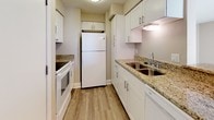 This single-floor residence offers a clean, modern aesthetic defined by warm neutral tones, white trim, and wood-look flooring that flows consistently throughout the space. The open-concept living and dining area is bright and welcoming, anchored by sliding glass doors fitted with vertical blinds that lead to a private balcony, filling the room with abundant natural light. A peninsula with granite countertops connects the living area to the galley-style kitchen, which is thoughtfully designed with white shaker cabinetry, granite countertops, a stainless steel sink, and white appliances including a refrigerator and range. Ample upper and lower cabinet storage maximizes the kitchen's functionality. The layout includes two well-sized bedrooms, each featuring vertical blind-covered windows that invite natural light. The two bathrooms are finished consistently with granite-topped white vanities and wood-look flooring. The full bathroom includes a tub and shower combination with tiled surround, while the second bathroom features a toilet and vanity. A closet accessible from one of the bathrooms provides additional storage. The overall ambiance of this space is bright, clean, and move-in ready, with cohesive finishes that lend a polished, contemporary feel throughout.