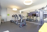 This single-level fitness room offers a well-organized and functional space for exercise, featuring a thoughtful layout that maximizes available floor area. The room is finished with durable dark rubber flooring suited for high-traffic athletic use, and the walls are painted in a clean, neutral tone that keeps the space feeling bright and open.

A large floor-to-ceiling mirror spans one wall, visually expanding the room while allowing users to monitor their form during workouts. Adjacent to the mirror, a striking panoramic cityscape mural adds a bold, urban design element that gives the space character and energy. Cardio equipment includes treadmills, elliptical trainers, a stationary cycle, and a multi-station cable machine, all from the Life Fitness brand. A yellow modular shelving unit provides convenient storage, and posted fitness room rules and instructional charts are displayed on the walls for user reference.

Overhead, circular pendant light fixtures provide warm, even illumination throughout the space. Natural light filters in through a large glazed door that opens directly to an outdoor area, offering ventilation and a visual connection to the exterior. The overall ambiance is clean, modern, and practical.