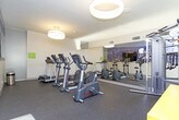 This single-level fitness room offers a well-organized and functional space for exercise, featuring a thoughtful layout that maximizes available floor area. The room is finished with durable dark rubber flooring suited for high-traffic athletic use, and the walls are painted in a clean, neutral tone that keeps the space feeling bright and open.

A large floor-to-ceiling mirror spans one wall, visually expanding the room while allowing users to monitor their form during workouts. Adjacent to the mirror, a striking panoramic cityscape mural adds a bold, urban design element that gives the space character and energy. Cardio equipment includes treadmills, elliptical trainers, a stationary cycle, and a multi-station cable machine, all from the Life Fitness brand. A yellow modular shelving unit provides convenient storage, and posted fitness room rules and instructional charts are displayed on the walls for user reference.

Overhead, circular pendant light fixtures provide warm, even illumination throughout the space. Natural light filters in through a large glazed door that opens directly to an outdoor area, offering ventilation and a visual connection to the exterior. The overall ambiance is clean, modern, and practical.