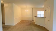 This single-floor space presents an open-concept layout that seamlessly connects the dining and living areas, creating a cohesive and functional flow. The dining area is anchored by an elegant chandelier that provides warm, ambient lighting and serves as a subtle focal point. A classic chair rail molding runs along the perimeter of the dining space, adding a touch of architectural detail to the otherwise clean, neutral walls painted in a soft off-white tone.

Plush, wall-to-wall carpeting in a warm beige hue extends throughout the space, contributing to a comfortable and inviting atmosphere. The transition toward the kitchen is defined by a curved bar-height counter with a dark countertop, offering a practical and stylish division between the living area and the kitchen beyond. This curved counter adds visual interest while maintaining the open feel of the overall layout.

A window on the far wall of the kitchen area allows natural light to filter into the space, brightening the interior. The overall design is neutral and move-in ready, with clean lines, minimal ornamentation, and versatile finishes that allow for a variety of interior design styles.