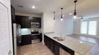 This single-floor space showcases a thoughtfully designed open-concept layout that seamlessly connects the kitchen to the living area. The kitchen is a standout feature, appointed with rich dark cabinetry that creates a striking contrast against the light granite countertops. A spacious kitchen island with a double stainless steel sink serves as the centerpiece, illuminated by three elegant pendant lights with clear glass shades. Stainless steel appliances, including a range and over-the-range microwave, add a polished, contemporary feel. A classic white subway tile backsplash completes the kitchen's refined aesthetic. Light-toned hardwood-style flooring runs throughout, lending warmth and continuity to the space. Recessed lighting is thoughtfully placed across the ceiling, ensuring the space is well-lit throughout. The open flow from the kitchen into the adjoining living area creates a sense of spaciousness, making the layout ideal for everyday living and entertaining. Neutral wall tones keep the overall ambiance clean, bright, and inviting, providing a versatile backdrop for a variety of interior styles.