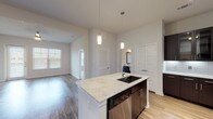 Located at 18610 Tuscany Stone in San Antonio, Texas, this single-floor residence offers a well-designed open-concept layout with modern finishes and thoughtful details throughout. The kitchen serves as the centerpiece of the living space, boasting a large granite-topped island with an undermount sink, dark espresso cabinetry, stainless steel appliances including a refrigerator, range, microwave, and dishwasher, complemented by a classic subway tile backsplash and elegant pendant lighting. Light wood-look flooring flows through the main living areas, while the open living room features elevated ceilings and a ceiling fan, creating an airy, spacious atmosphere. The bedroom is carpeted for comfort, includes a ceiling fan, and benefits from natural light through a window with blinds. The bathroom is appointed with a granite-topped dark vanity, a large framed mirror, and tile flooring, with direct access to a walk-in closet featuring built-in shelving. A dedicated laundry room accommodates a full-size washer and dryer. Throughout, the neutral color palette and clean architectural lines create a cohesive, contemporary ambiance ideal for modern living in the Stone Oak area of San Antonio.